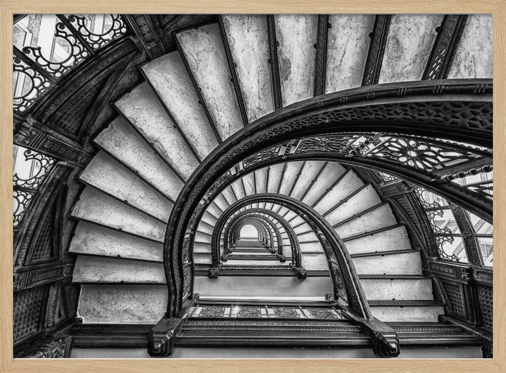 A dramatic black and white photograph looking up from the bottom of an ornate spiral staircase. The dark, intricate ironwork of the railings contrasts with the light stone steps, creating a mesmerizing, repeating pattern that spirals towards a bright light at the top. The image is presented within a silver-colored frame. Decor