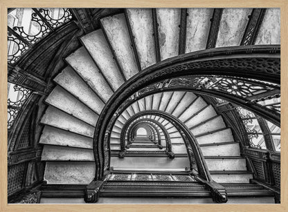 A dramatic black and white photograph looking up from the bottom of an ornate spiral staircase. The dark, intricate ironwork of the railings contrasts with the light stone steps, creating a mesmerizing, repeating pattern that spirals towards a bright light at the top. The image is presented within a silver-colored frame. Decor