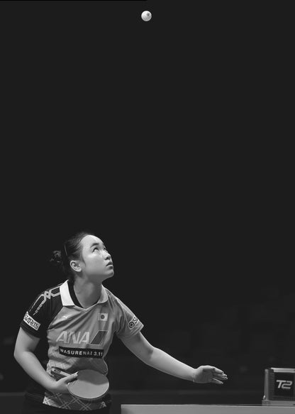 A dramatic black and white vertical photograph of a female table tennis player mid-serve. Positioned in the lower-left, she looks up with intense focus at the white ball suspended high in the air against a solid black background. Poster