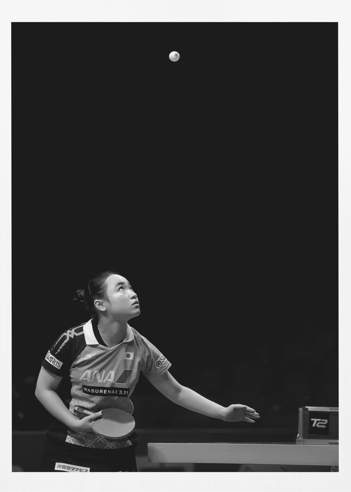 A dramatic black and white vertical photograph of a female table tennis player mid-serve. Positioned in the lower-left, she looks up with intense focus at the white ball suspended high in the air against a solid black background. Poster