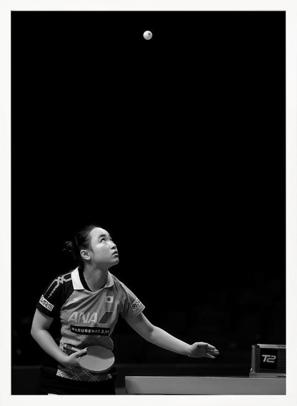 A dramatic black and white vertical photograph of a female table tennis player mid-serve. Positioned in the lower-left, she looks up with intense focus at the white ball suspended high in the air against a solid black background. Poster