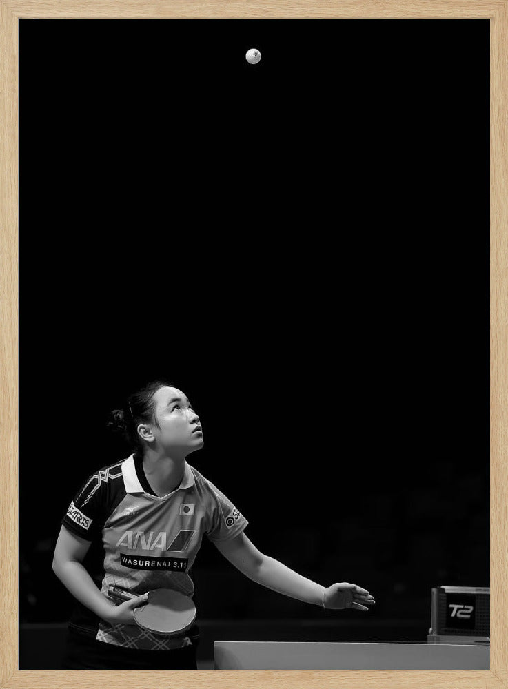 A dramatic black and white vertical photograph of a female table tennis player mid-serve. Positioned in the lower-left, she looks up with intense focus at the white ball suspended high in the air against a solid black background. Poster