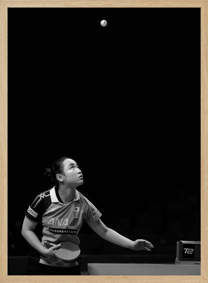 A dramatic black and white vertical photograph of a female table tennis player mid-serve. Positioned in the lower-left, she looks up with intense focus at the white ball suspended high in the air against a solid black background. Poster