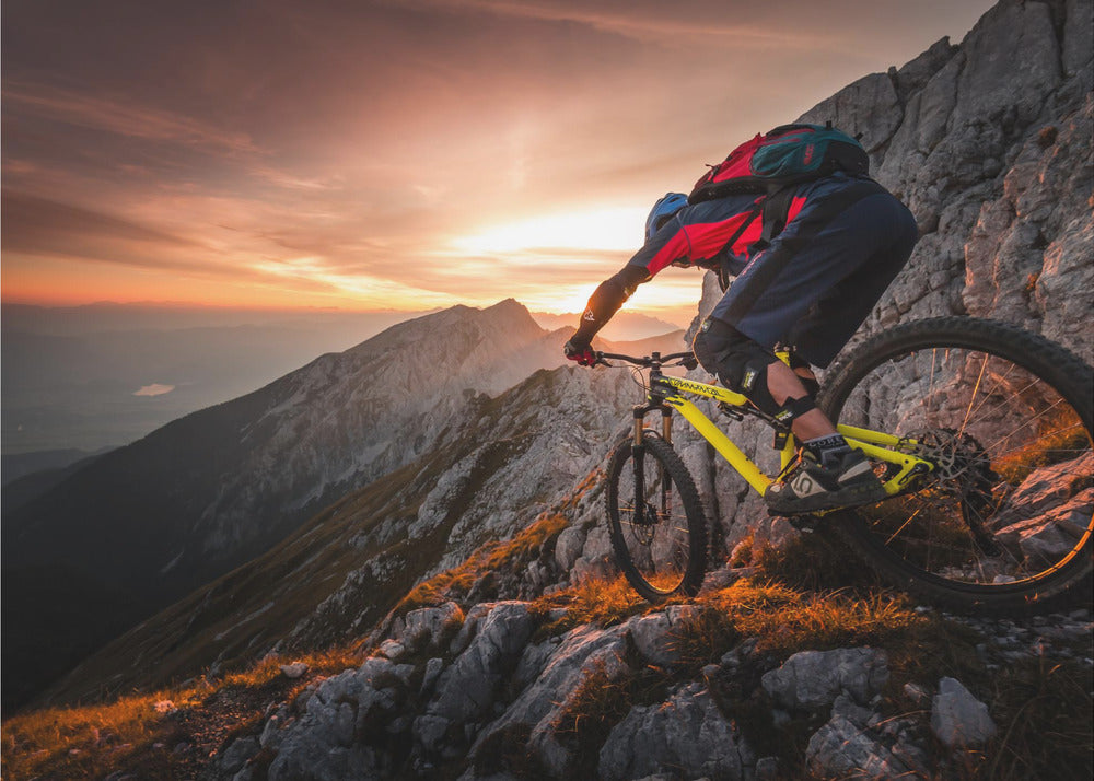A mountain biker in full gear rides a yellow bike down a narrow, rocky mountain ridge at sunset. The sky is filled with the warm colors of the setting sun, which illuminates the distant mountain peaks. Artwork