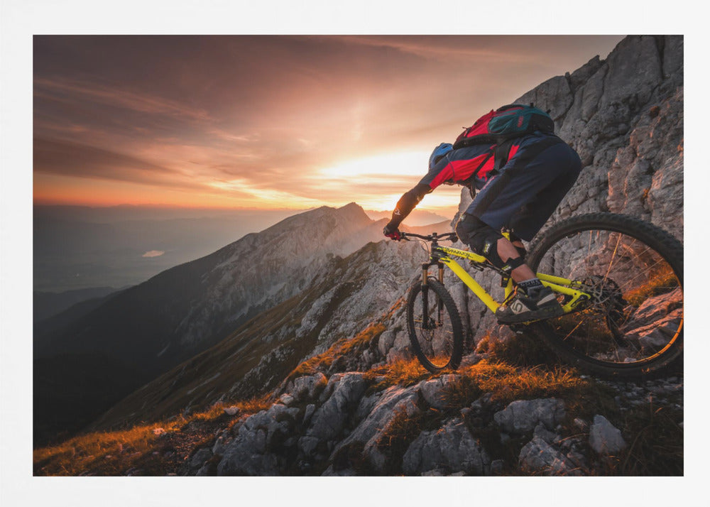A mountain biker in full gear rides a yellow bike down a narrow, rocky mountain ridge at sunset. The sky is filled with the warm colors of the setting sun, which illuminates the distant mountain peaks. Artwork