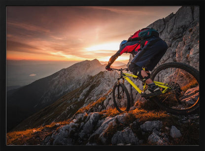 A mountain biker in full gear rides a yellow bike down a narrow, rocky mountain ridge at sunset. The sky is filled with the warm colors of the setting sun, which illuminates the distant mountain peaks. Artwork
