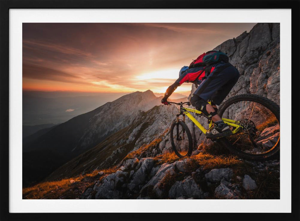 A mountain biker in full gear rides a yellow bike down a narrow, rocky mountain ridge at sunset. The sky is filled with the warm colors of the setting sun, which illuminates the distant mountain peaks. Artwork