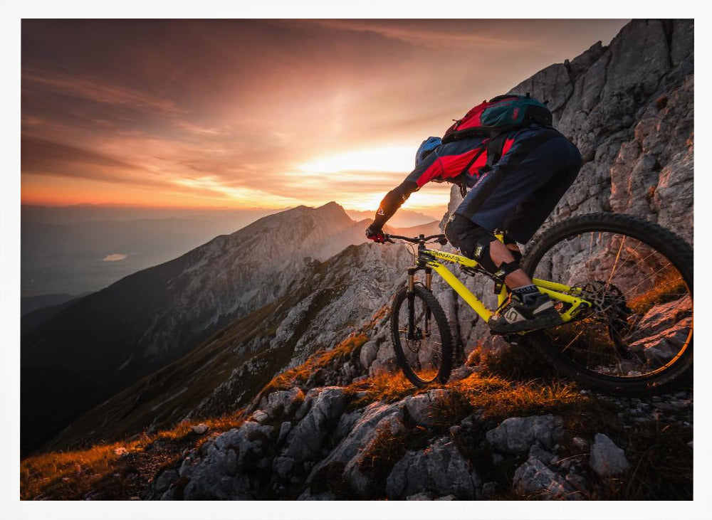 A mountain biker in full gear rides a yellow bike down a narrow, rocky mountain ridge at sunset. The sky is filled with the warm colors of the setting sun, which illuminates the distant mountain peaks. Artwork
