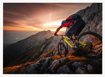 A mountain biker in full gear rides a yellow bike down a narrow, rocky mountain ridge at sunset. The sky is filled with the warm colors of the setting sun, which illuminates the distant mountain peaks. Artwork