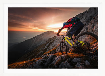 A mountain biker in full gear rides a yellow bike down a narrow, rocky mountain ridge at sunset. The sky is filled with the warm colors of the setting sun, which illuminates the distant mountain peaks. Artwork