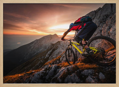 A mountain biker in full gear rides a yellow bike down a narrow, rocky mountain ridge at sunset. The sky is filled with the warm colors of the setting sun, which illuminates the distant mountain peaks. Artwork