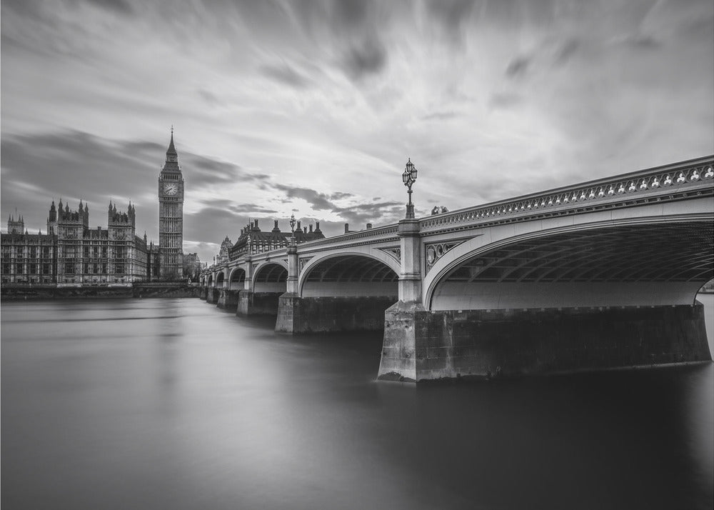 A dramatic black and white long-exposure photograph of London, featuring the smooth River Thames in the foreground, Westminster Bridge on the right, and the iconic Big Ben and Houses of Parliament on the left under a cloudy, streaked sky. The image is presented in a silver frame. Print
