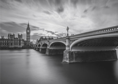 A dramatic black and white long-exposure photograph of London, featuring the smooth River Thames in the foreground, Westminster Bridge on the right, and the iconic Big Ben and Houses of Parliament on the left under a cloudy, streaked sky. The image is presented in a silver frame. Print