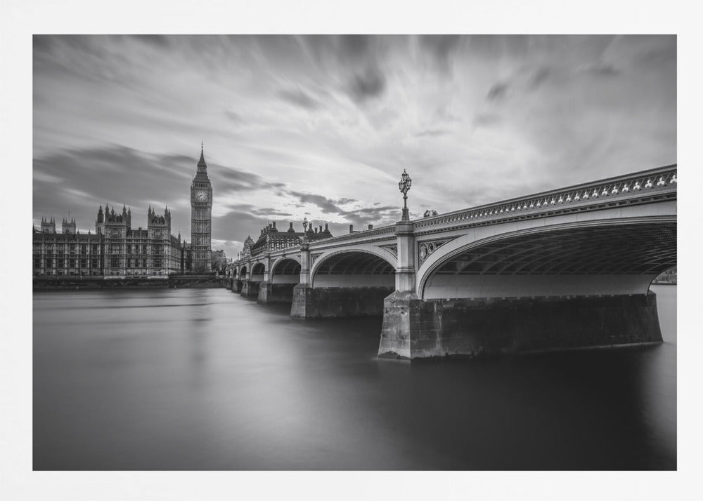 A dramatic black and white long-exposure photograph of London, featuring the smooth River Thames in the foreground, Westminster Bridge on the right, and the iconic Big Ben and Houses of Parliament on the left under a cloudy, streaked sky. The image is presented in a silver frame. Print