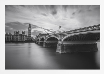 A dramatic black and white long-exposure photograph of London, featuring the smooth River Thames in the foreground, Westminster Bridge on the right, and the iconic Big Ben and Houses of Parliament on the left under a cloudy, streaked sky. The image is presented in a silver frame. Print