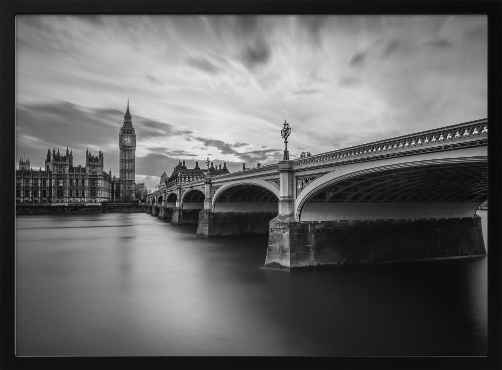 A dramatic black and white long-exposure photograph of London, featuring the smooth River Thames in the foreground, Westminster Bridge on the right, and the iconic Big Ben and Houses of Parliament on the left under a cloudy, streaked sky. The image is presented in a silver frame. Print