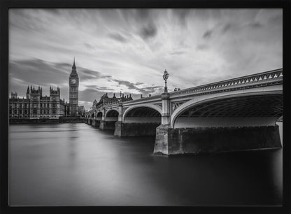 A dramatic black and white long-exposure photograph of London, featuring the smooth River Thames in the foreground, Westminster Bridge on the right, and the iconic Big Ben and Houses of Parliament on the left under a cloudy, streaked sky. The image is presented in a silver frame. Print