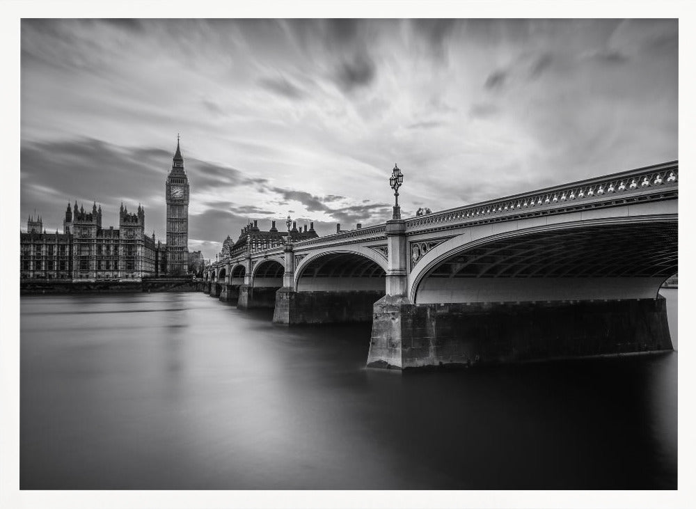 A dramatic black and white long-exposure photograph of London, featuring the smooth River Thames in the foreground, Westminster Bridge on the right, and the iconic Big Ben and Houses of Parliament on the left under a cloudy, streaked sky. The image is presented in a silver frame. Print