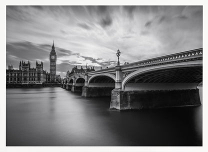 A dramatic black and white long-exposure photograph of London, featuring the smooth River Thames in the foreground, Westminster Bridge on the right, and the iconic Big Ben and Houses of Parliament on the left under a cloudy, streaked sky. The image is presented in a silver frame. Print