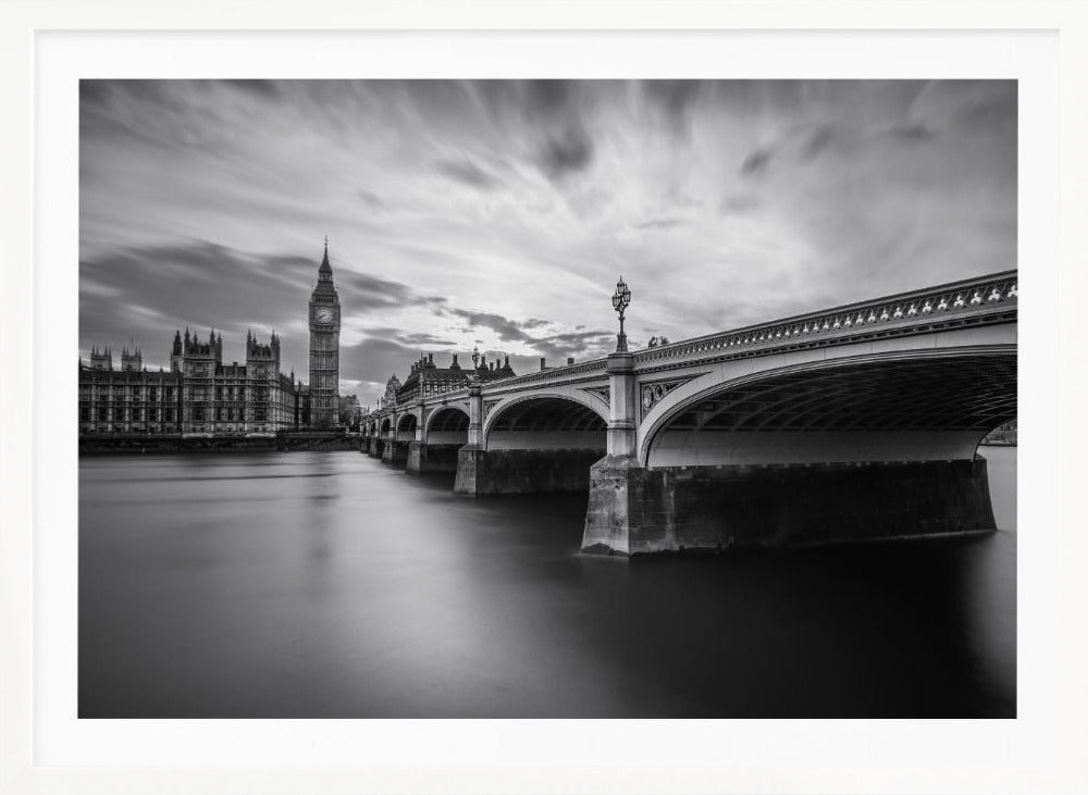 A dramatic black and white long-exposure photograph of London, featuring the smooth River Thames in the foreground, Westminster Bridge on the right, and the iconic Big Ben and Houses of Parliament on the left under a cloudy, streaked sky. The image is presented in a silver frame. Print