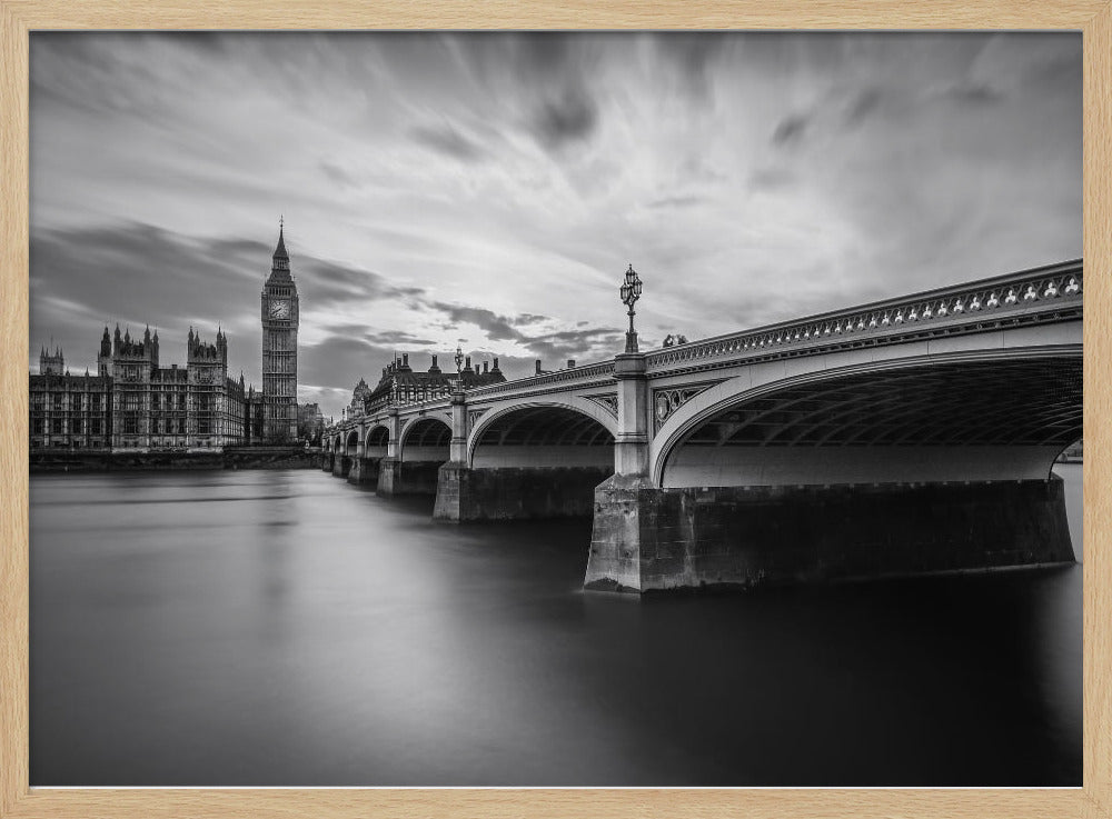 A dramatic black and white long-exposure photograph of London, featuring the smooth River Thames in the foreground, Westminster Bridge on the right, and the iconic Big Ben and Houses of Parliament on the left under a cloudy, streaked sky. The image is presented in a silver frame. Print