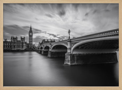 A dramatic black and white long-exposure photograph of London, featuring the smooth River Thames in the foreground, Westminster Bridge on the right, and the iconic Big Ben and Houses of Parliament on the left under a cloudy, streaked sky. The image is presented in a silver frame. Print