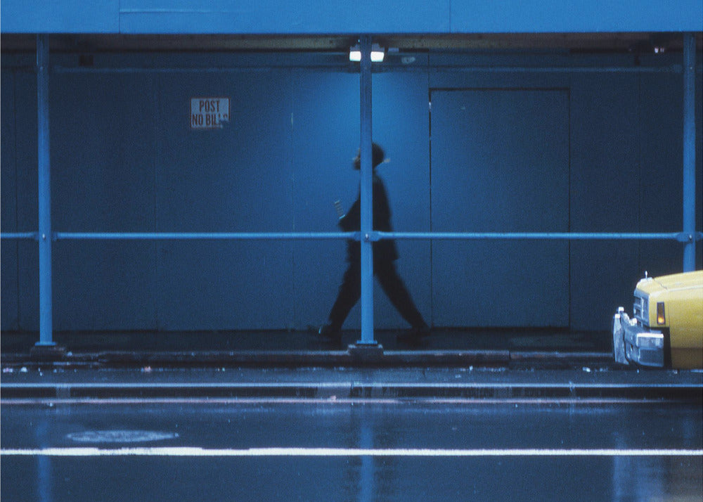 A moody, grainy photograph dominated by shades of blue, capturing the blurred silhouette of a person walking under a covered city sidewalk at night. The wet street in the foreground reflects light, and the yellow cab of a truck is partially visible on the right. Artwork