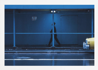 A moody, grainy photograph dominated by shades of blue, capturing the blurred silhouette of a person walking under a covered city sidewalk at night. The wet street in the foreground reflects light, and the yellow cab of a truck is partially visible on the right. Artwork