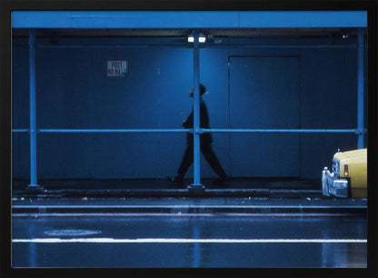 A moody, grainy photograph dominated by shades of blue, capturing the blurred silhouette of a person walking under a covered city sidewalk at night. The wet street in the foreground reflects light, and the yellow cab of a truck is partially visible on the right. Artwork