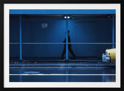 A moody, grainy photograph dominated by shades of blue, capturing the blurred silhouette of a person walking under a covered city sidewalk at night. The wet street in the foreground reflects light, and the yellow cab of a truck is partially visible on the right. Artwork