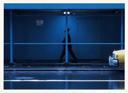 A moody, grainy photograph dominated by shades of blue, capturing the blurred silhouette of a person walking under a covered city sidewalk at night. The wet street in the foreground reflects light, and the yellow cab of a truck is partially visible on the right. Artwork
