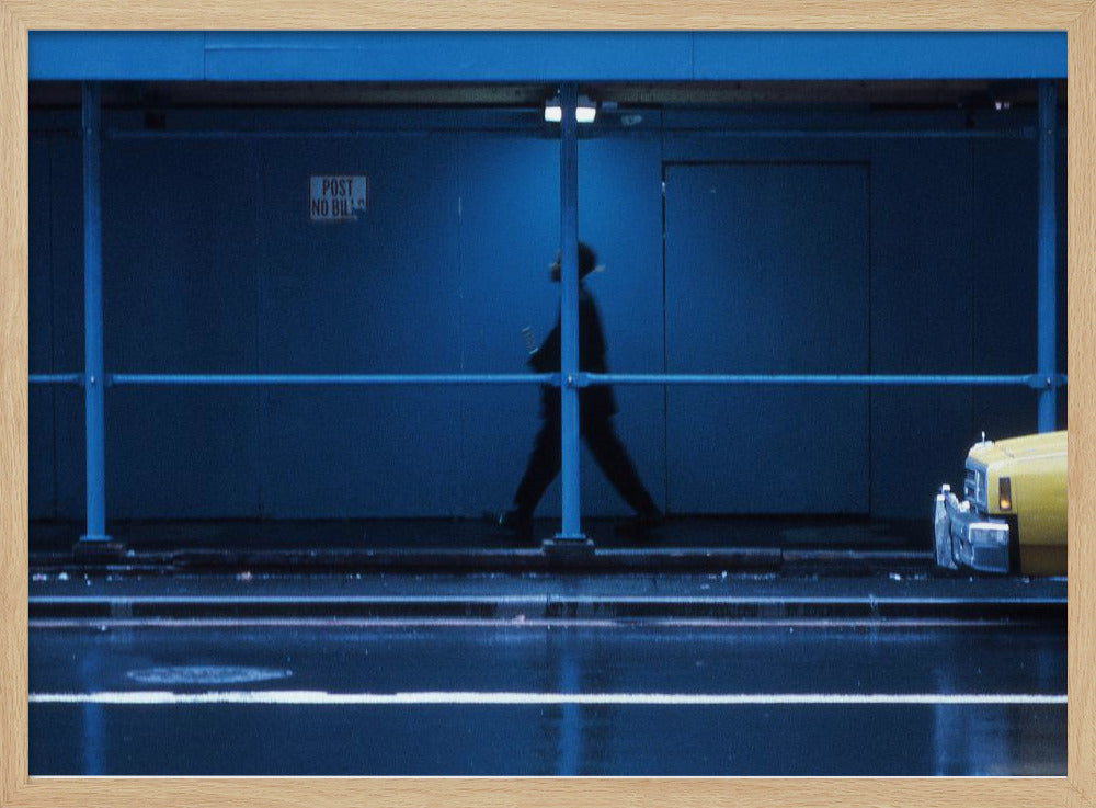 A moody, grainy photograph dominated by shades of blue, capturing the blurred silhouette of a person walking under a covered city sidewalk at night. The wet street in the foreground reflects light, and the yellow cab of a truck is partially visible on the right. Artwork