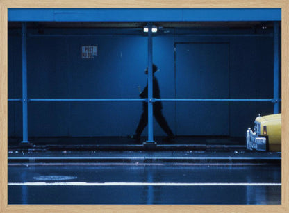 A moody, grainy photograph dominated by shades of blue, capturing the blurred silhouette of a person walking under a covered city sidewalk at night. The wet street in the foreground reflects light, and the yellow cab of a truck is partially visible on the right. Artwork