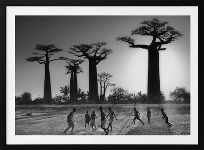 A framed, black and white photograph capturing a vibrant scene of several children playing soccer on a dirt field. In the background, the iconic silhouettes of large Baobab trees stand tall against a bright, hazy sun, casting long shadows of the players across the ground. Artwork