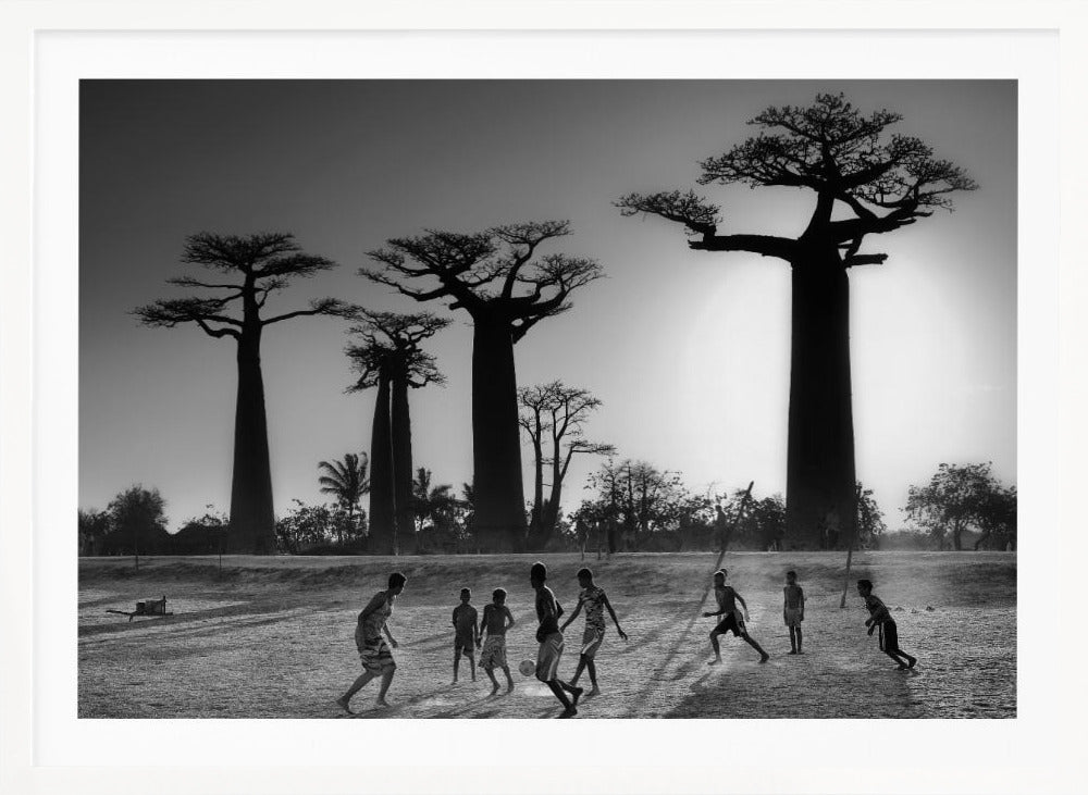 A framed, black and white photograph capturing a vibrant scene of several children playing soccer on a dirt field. In the background, the iconic silhouettes of large Baobab trees stand tall against a bright, hazy sun, casting long shadows of the players across the ground. Artwork