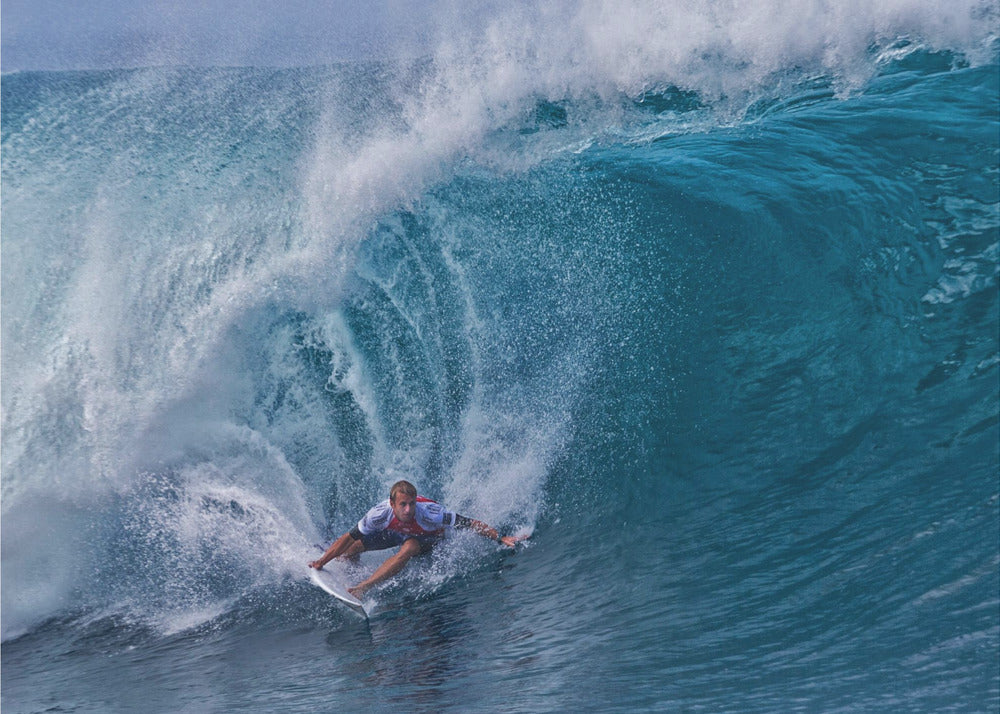 A dramatic action shot of a professional surfer skillfully navigating the inside of a massive, curling blue ocean wave, surrounded by white sea spray, all enclosed in a silver frame. Artwork