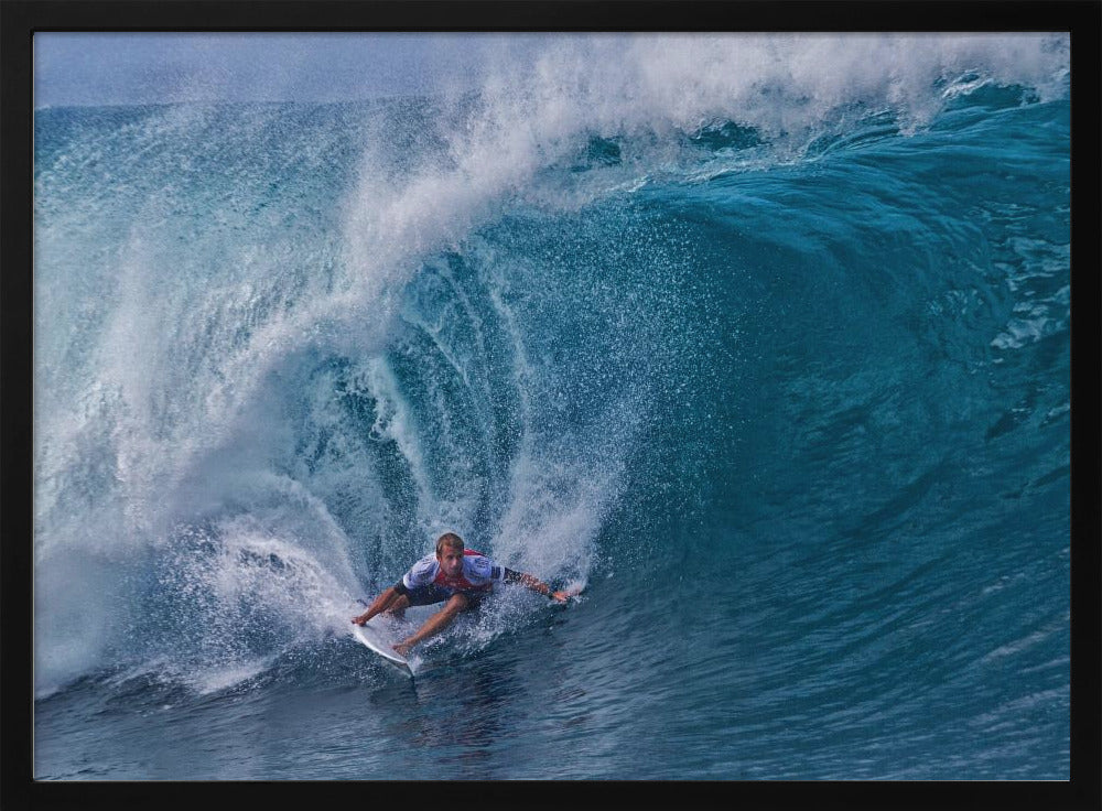A dramatic action shot of a professional surfer skillfully navigating the inside of a massive, curling blue ocean wave, surrounded by white sea spray, all enclosed in a silver frame. Artwork