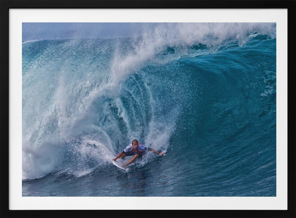 A dramatic action shot of a professional surfer skillfully navigating the inside of a massive, curling blue ocean wave, surrounded by white sea spray, all enclosed in a silver frame. Artwork