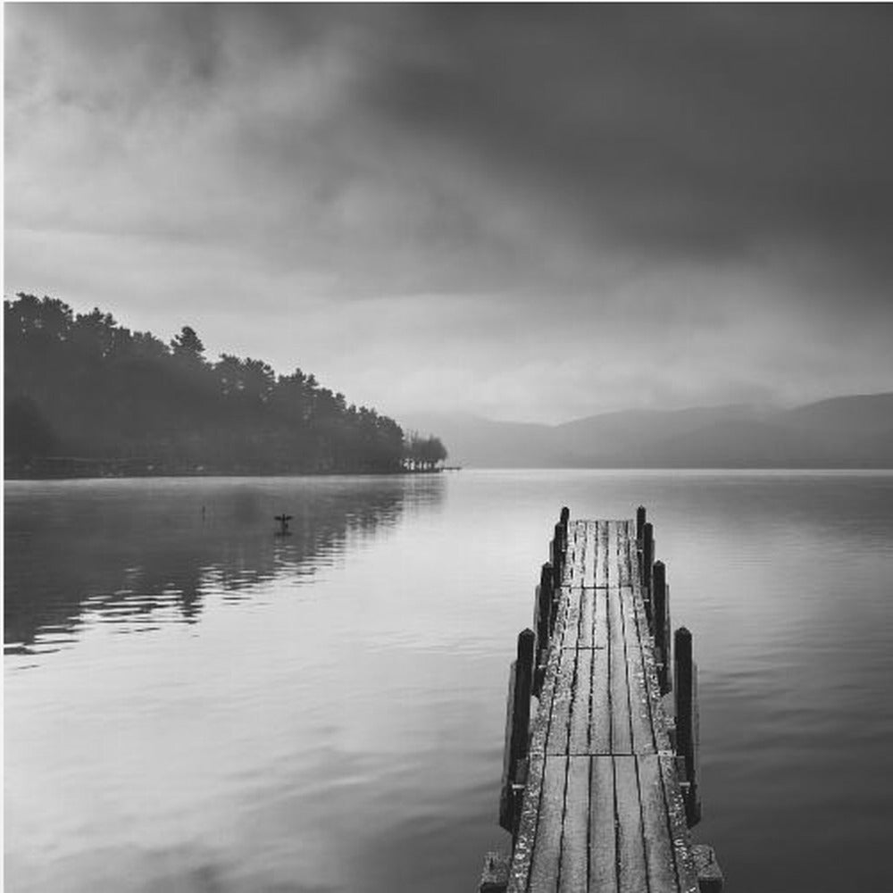 A framed, moody black and white photograph of a wooden pier extending into a calm lake under a cloudy sky, with a forested shoreline and distant mountains in the background. Poster