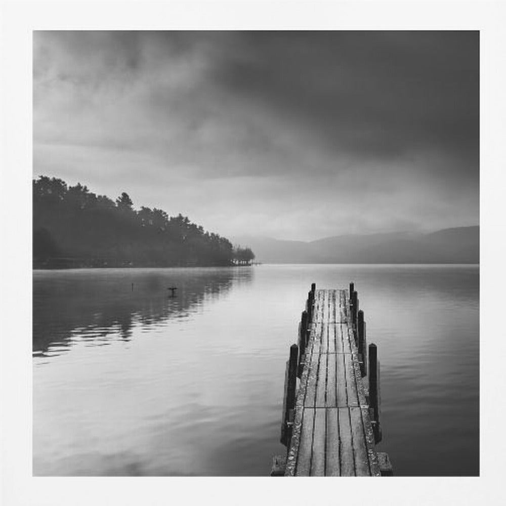 A framed, moody black and white photograph of a wooden pier extending into a calm lake under a cloudy sky, with a forested shoreline and distant mountains in the background. Poster