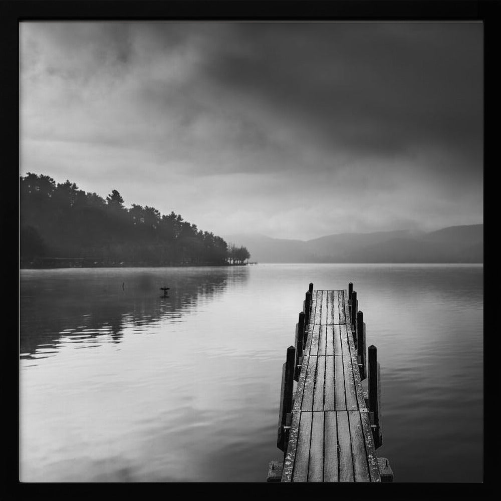 A framed, moody black and white photograph of a wooden pier extending into a calm lake under a cloudy sky, with a forested shoreline and distant mountains in the background. Poster