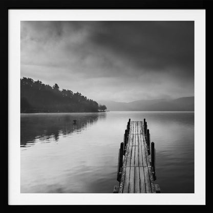A framed, moody black and white photograph of a wooden pier extending into a calm lake under a cloudy sky, with a forested shoreline and distant mountains in the background. Poster