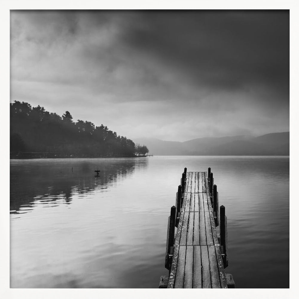 A framed, moody black and white photograph of a wooden pier extending into a calm lake under a cloudy sky, with a forested shoreline and distant mountains in the background. Poster