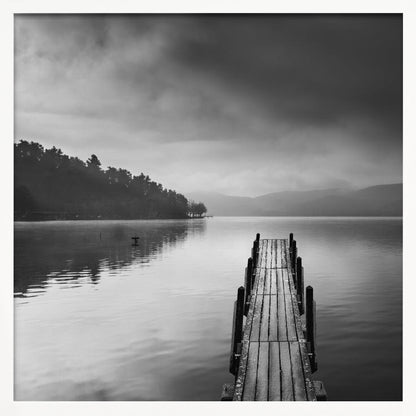 A framed, moody black and white photograph of a wooden pier extending into a calm lake under a cloudy sky, with a forested shoreline and distant mountains in the background. Poster