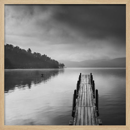 A framed, moody black and white photograph of a wooden pier extending into a calm lake under a cloudy sky, with a forested shoreline and distant mountains in the background. Poster