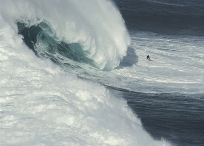 A framed aerial photograph captures a dramatic ocean scene where a lone surfer on a surfboard is dwarfed by an immense, powerful wave cresting nearby, conveying a sense of scale and adventure. Artwork
