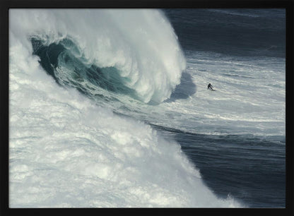 A framed aerial photograph captures a dramatic ocean scene where a lone surfer on a surfboard is dwarfed by an immense, powerful wave cresting nearby, conveying a sense of scale and adventure. Artwork