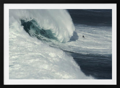 A framed aerial photograph captures a dramatic ocean scene where a lone surfer on a surfboard is dwarfed by an immense, powerful wave cresting nearby, conveying a sense of scale and adventure. Artwork