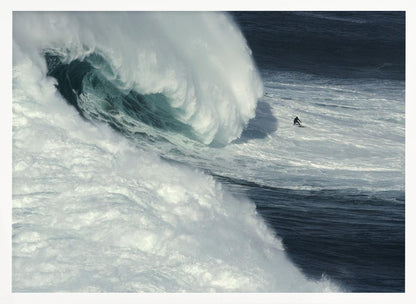 A framed aerial photograph captures a dramatic ocean scene where a lone surfer on a surfboard is dwarfed by an immense, powerful wave cresting nearby, conveying a sense of scale and adventure. Artwork