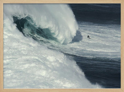 A framed aerial photograph captures a dramatic ocean scene where a lone surfer on a surfboard is dwarfed by an immense, powerful wave cresting nearby, conveying a sense of scale and adventure. Artwork
